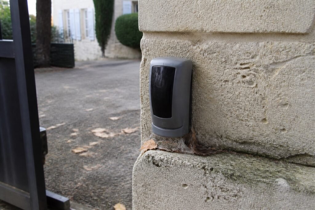Cellule photoélectrique fixée à faible hauteur sur le pilier d'un portail, avec des feuilles mortes au pied